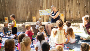 Teacher reads picture book to children sitting on blankets in a sunny fenced backyard, with cups and a calm, attentive mood.