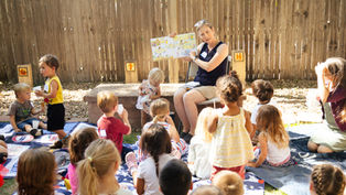 Teacher reads picture book to children sitting on blankets in a sunny fenced backyard, with cups and a calm, attentive mood.