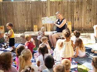 Teacher reads picture book to children sitting on blankets in a sunny fenced backyard, with cups and a calm, attentive mood.