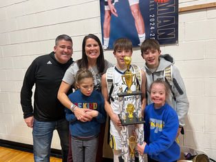 A group of smiling people poses with a sports trophy in a gym. Two individuals wear "BE KIND" shirts. A poster hangs in the background.