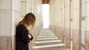 A woman in a patterned dress leans against a wall in a sunlit corridor, gazing downwards, evoking a contemplative mood.