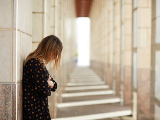 A woman in a patterned dress leans against a wall in a sunlit corridor, gazing downwards, evoking a contemplative mood.