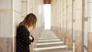 A woman in a patterned dress leans against a wall in a sunlit corridor, gazing downwards, evoking a contemplative mood.