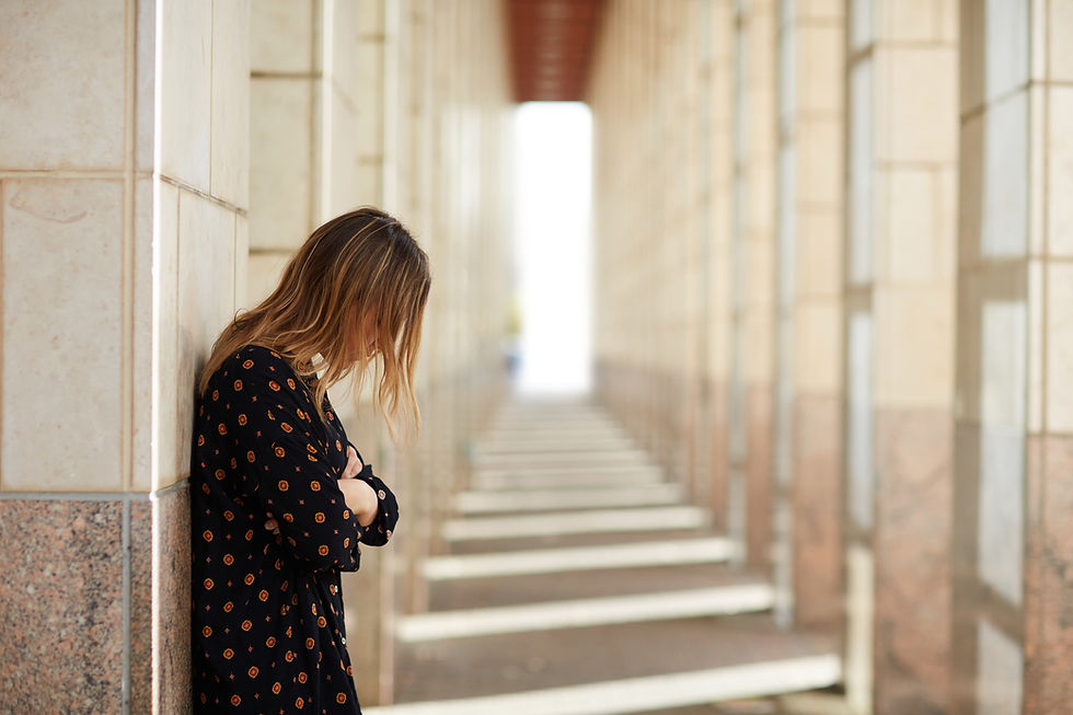 A woman in a patterned dress leans against a wall in a sunlit corridor, gazing downwards, evoking a contemplative mood.