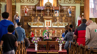 A congregation stands in pews during a church service. A priest in green stands at an ornate altar with statues and colorful flowers.