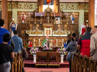 A congregation stands in pews during a church service. A priest in green stands at an ornate altar with statues and colorful flowers.