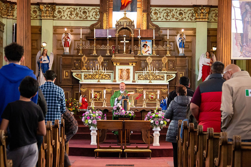 A congregation stands in pews during a church service. A priest in green stands at an ornate altar with statues and colorful flowers.