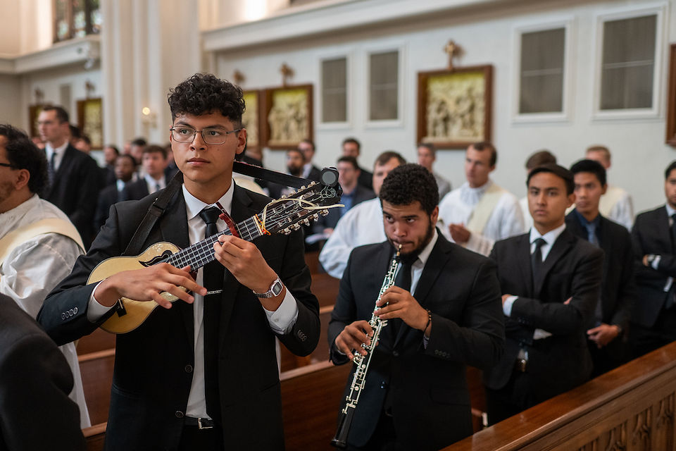 Men in suits play guitar and oboe during a ceremony in a church. Others in suits and robes stand behind them, creating a formal ambiance.