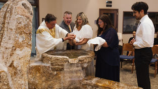 Priest baptizes a baby at a stone baptismal font while family and friends watch in a church.