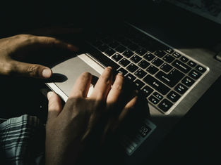 Hands typing on a laptop keyboard in dim light. Shadows and a plaid pattern in the lower left create a focused, moody atmosphere.