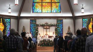 Church service with congregants seated, priests at altar under a crucifix. Colorful stained glass windows and floral decor create reverence.
