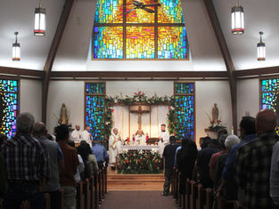 Church service with congregants seated, priests at altar under a crucifix. Colorful stained glass windows and floral decor create reverence.