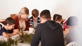 Family of six holds hands, heads bowed in prayer around a table with greenery and brass items. Warm, cozy ambiance.