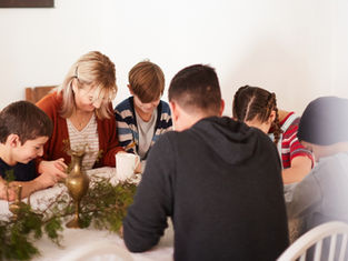 Family of six holds hands, heads bowed in prayer around a table with greenery and brass items. Warm, cozy ambiance.