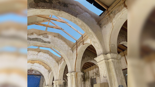 Weathered stone arches in a partially roofless building under renovation. Visible blue sky and wooden beams. Text: Photo: Bishop Cisneros.