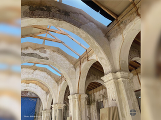 Weathered stone arches in a partially roofless building under renovation. Visible blue sky and wooden beams. Text: Photo: Bishop Cisneros.