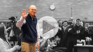 Elderly man waves, smiling, in a colorful vest amid monochrome crowd applauding. Brick wall and clock in background. Play button overlay.