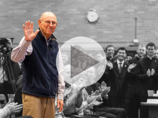 Elderly man waves, smiling, in a colorful vest amid monochrome crowd applauding. Brick wall and clock in background. Play button overlay.