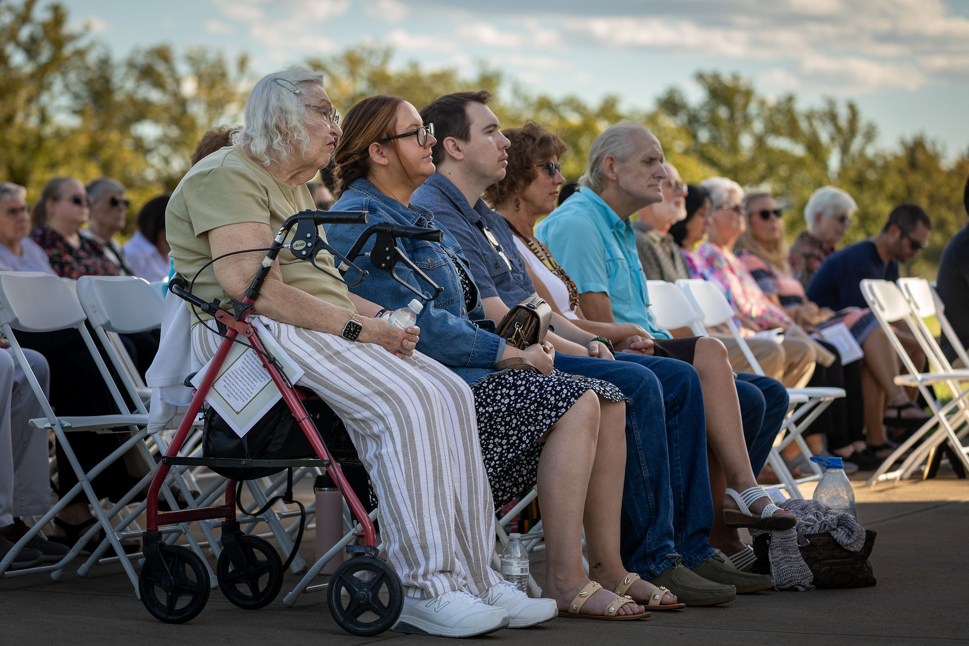 People seated outdoors in folding chairs, attentively listening. A woman with a walker sits beside others. Greenery in the background, clear sky.