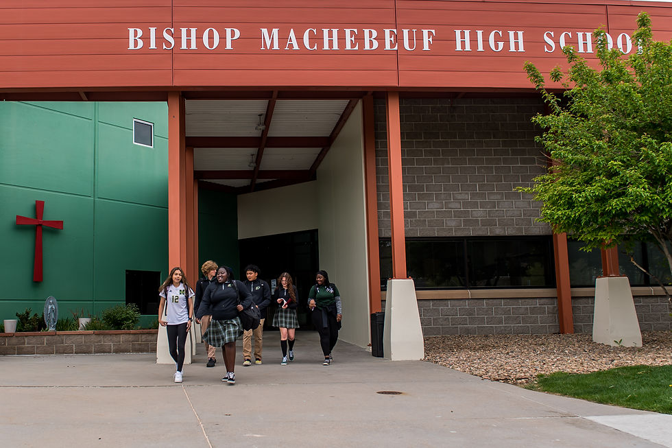 Students walk in front of Bishop Machebeuf High School, set against a red and green facade with a cross. A tree adds greenery.