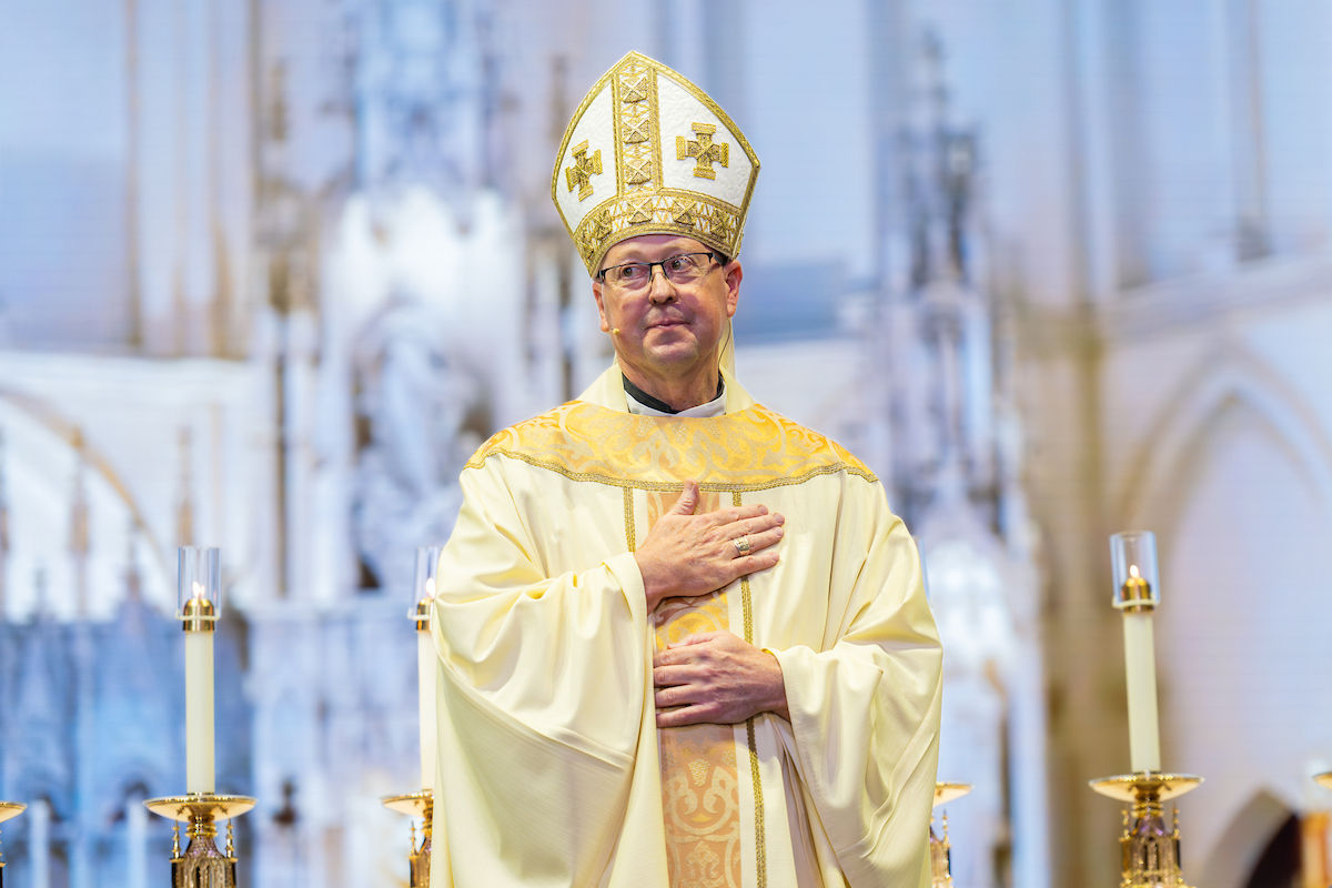 Bishop in ornate vestments stands before a cathedral backdrop, hand on chest. Four candles frame the scene, evoking a solemn mood.