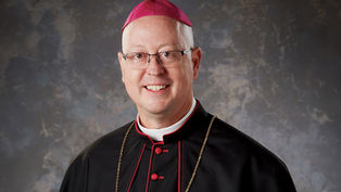 Smiling man in clerical dress with a magenta zucchetto and gold chain against a textured gray background.