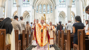 Bishop in ornate robes and mitre gestures during church service procession in cathedral with stained glass windows, surrounded by congregation.