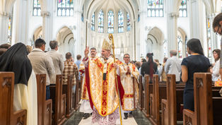 Bishop in ornate robes and mitre gestures during church service procession in cathedral with stained glass windows, surrounded by congregation.