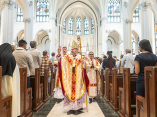Bishop in ornate robes and mitre gestures during church service procession in cathedral with stained glass windows, surrounded by congregation.