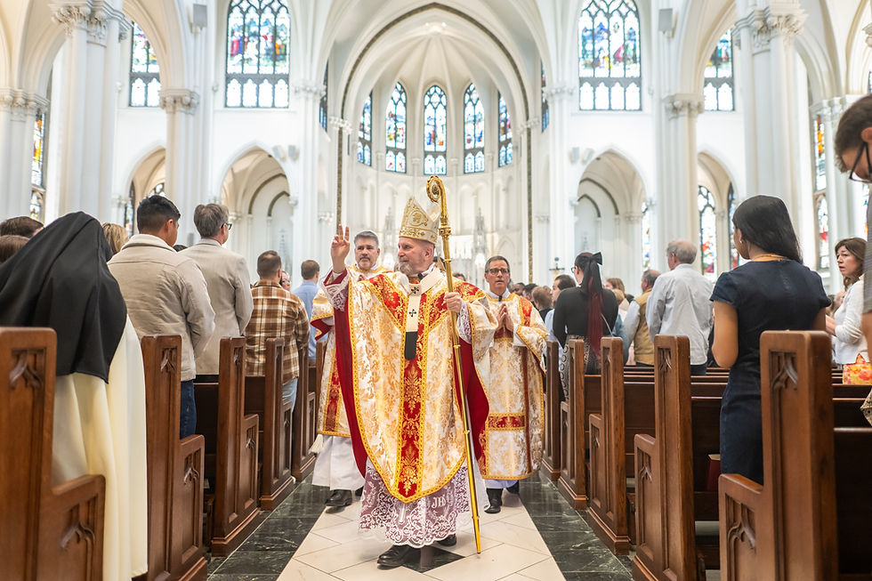 Bishop in ornate robes and mitre gestures during church service procession in cathedral with stained glass windows, surrounded by congregation.
