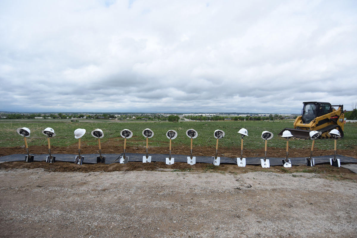 Hard hats on shovels lined up in a field under cloudy skies, with a yellow bulldozer in the background, suggesting a groundbreaking event.