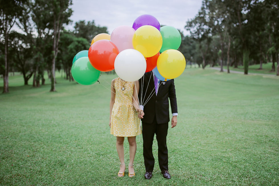 Couple in a field holding colorful balloons, obscuring faces. Woman in yellow dress, man in suit. Green grass and trees in background.
