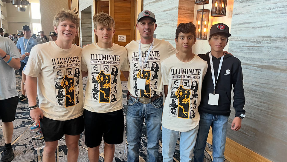 Five people stand together wearing "Illuminate Steubenville Conferences" shirts. They are indoors, with a wooden door and lamps in the background.
