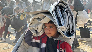A boy carries a folded tent on his head in a crowded setting. People, some with bags, walk beside him. Dusty environment, somber mood.