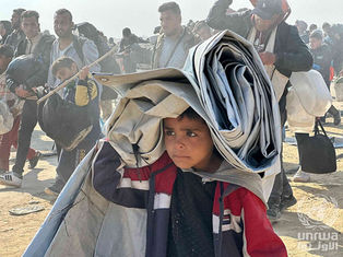 A boy carries a folded tent on his head in a crowded setting. People, some with bags, walk beside him. Dusty environment, somber mood.