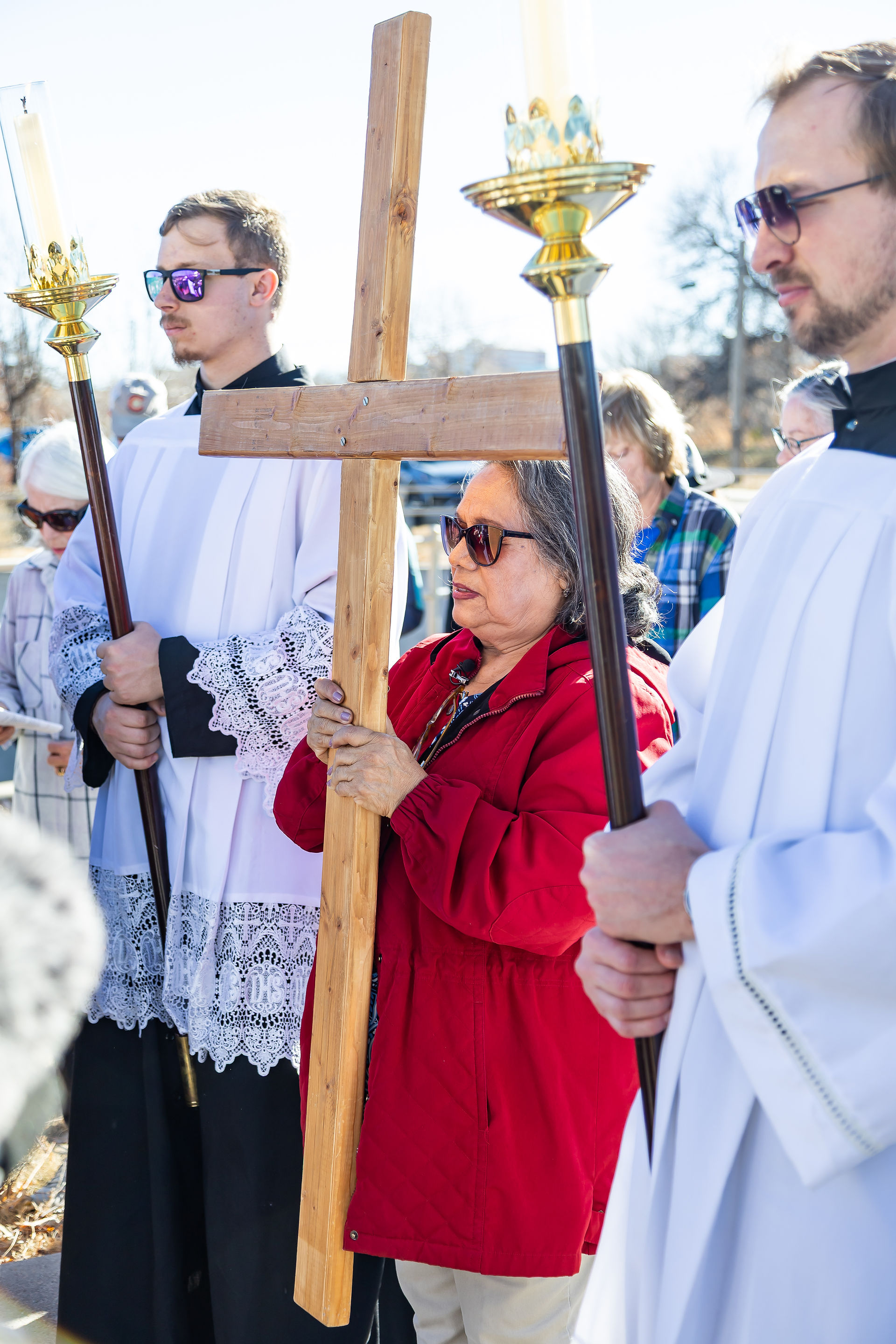 A woman in a red jacket holds a wooden cross, flanked by two men in white robes holding candles. Outdoor setting with clear skies.