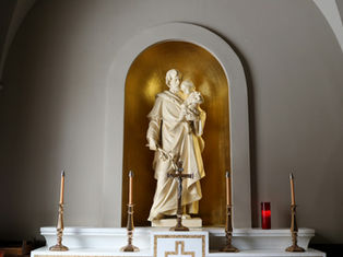 Statue of a robed figure holding a child in a gold alcove, surrounded by four candlesticks and a crucifix on a white altar. Calm mood.