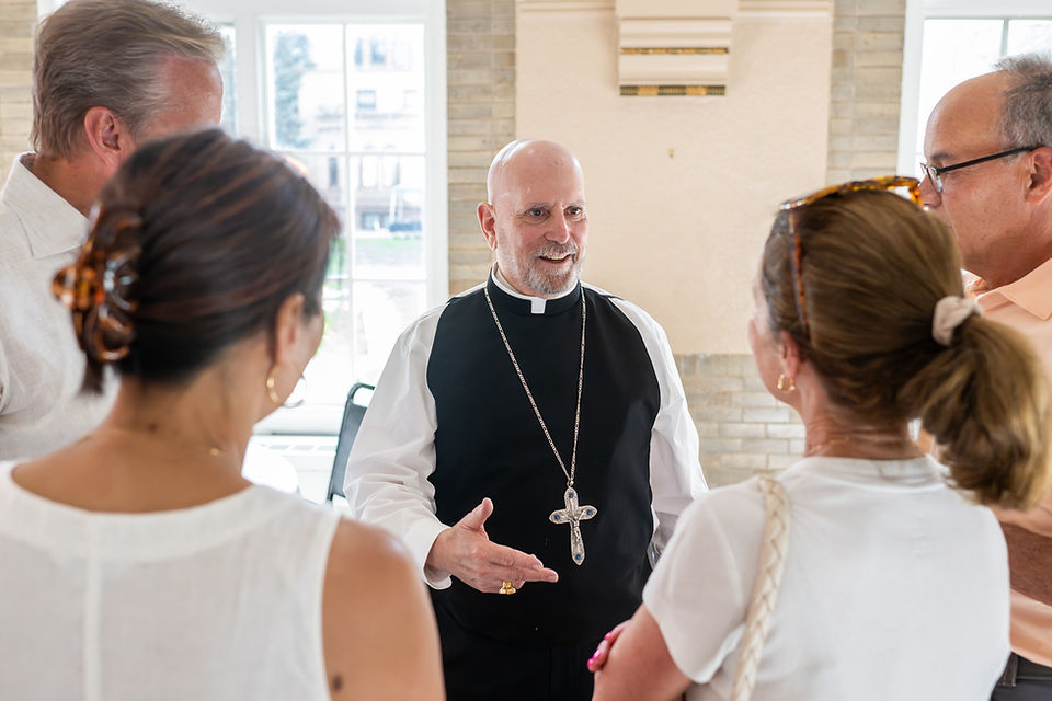 Priest with large cross speaks to a small group indoors, smiling in a bright room with windows.