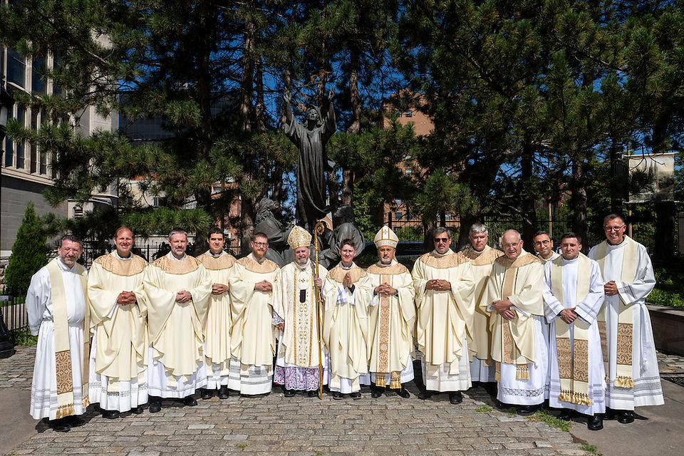 A group of clergy in white robes and gold stoles stand outside among trees, posing for a photo on a sunny day. A statue is in the background.