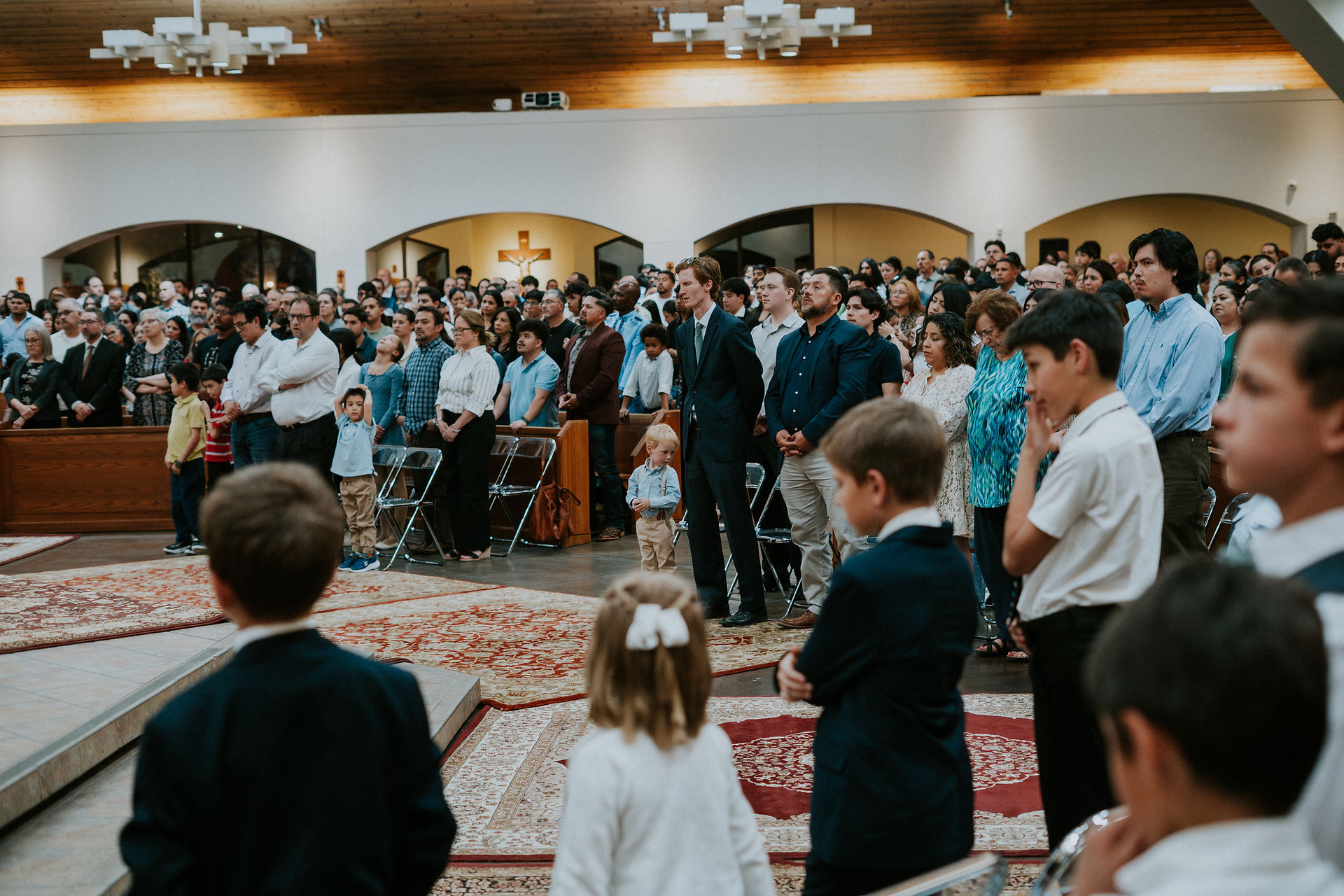 A congregation stands in a church, facing front. The setting is calm, with wooden ceilings and patterned rugs. People wear formal attire.