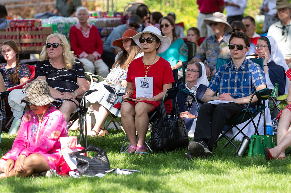 Crowd seated on grass at an outdoor gathering, many in hats and sunglasses, including nuns and a woman in red holding a pamphlet.