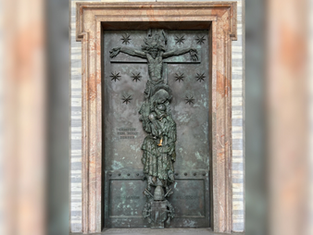 Bronze relief on a door showing a figure on a cross with stars. Text reads "CHRISTUS HER MUNDI" and dates "A.D. 2000." Stone frame.