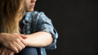 Woman in a blue polka-dot shirt, sitting with hands clasped, gazes thoughtfully to the side against a dark background.