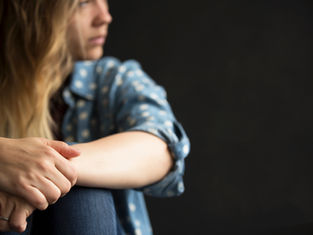 Woman in a blue polka-dot shirt, sitting with hands clasped, gazes thoughtfully to the side against a dark background.