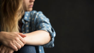 Woman in a blue polka-dot shirt, sitting with hands clasped, gazes thoughtfully to the side against a dark background.