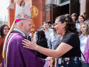 A person in a purple robe and mitre greets a smiling woman outside a brick building, surrounded by a group of people. Festive and friendly mood.