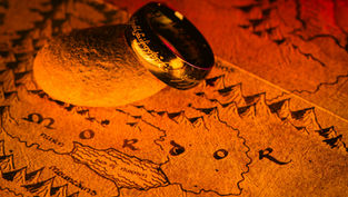 A golden ring with inscriptions lies on a rock atop a sepia-toned map of Mordor, in warm, dramatic lighting.