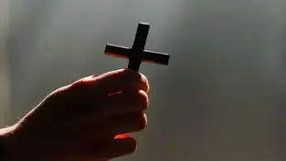 A hand holds a small cross against a dimly lit background, creating a solemn, reflective mood. Light casts a gentle glow on the fingers.