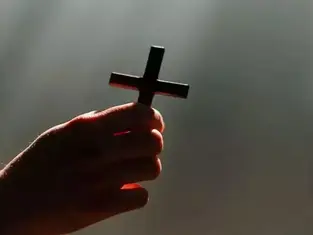 A hand holds a small cross against a dimly lit background, creating a solemn, reflective mood. Light casts a gentle glow on the fingers.