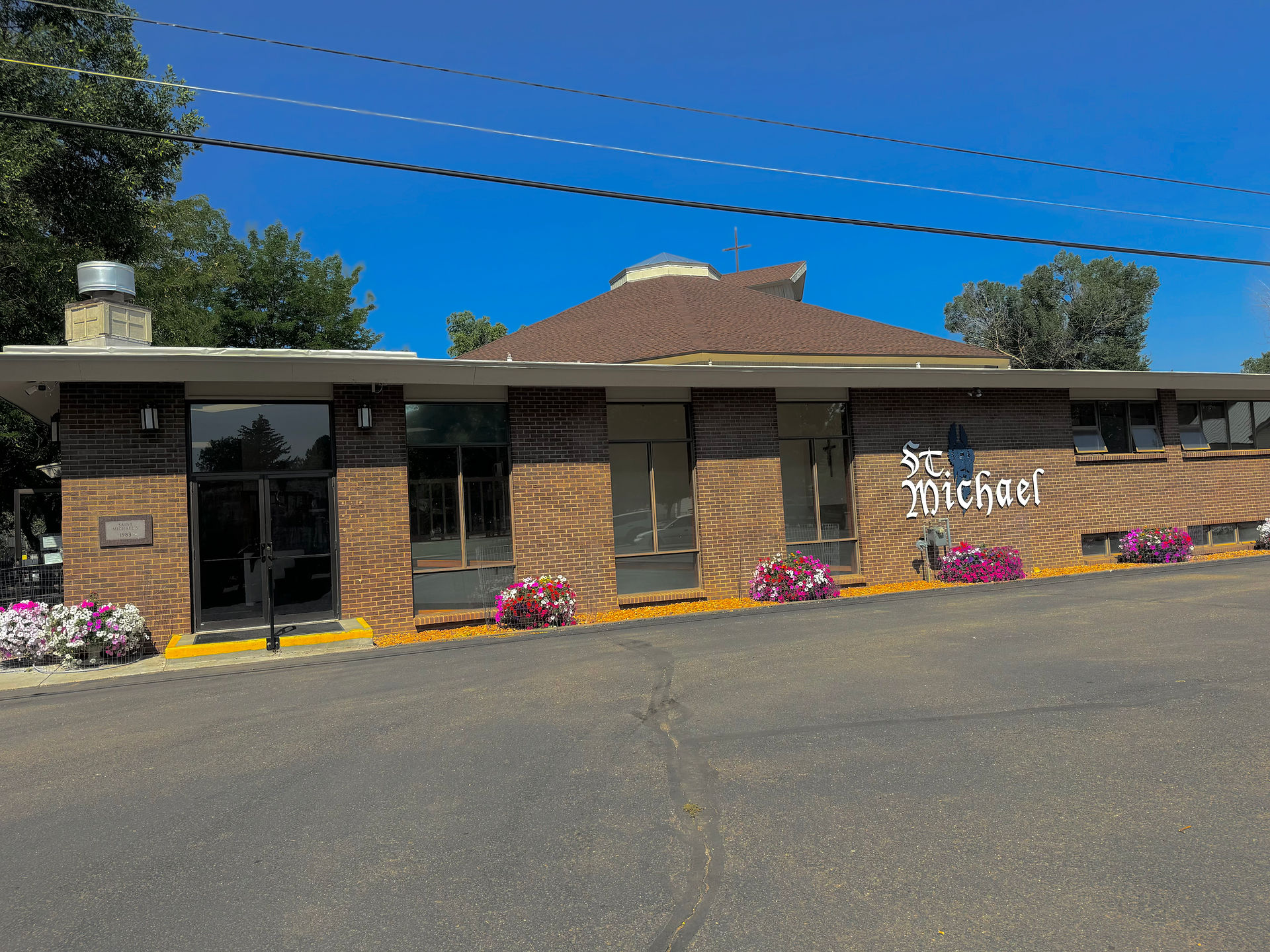 Brick building with "St. Michael" sign, surrounded by colorful flowers. Clear blue sky with trees in the background. Calm atmosphere.